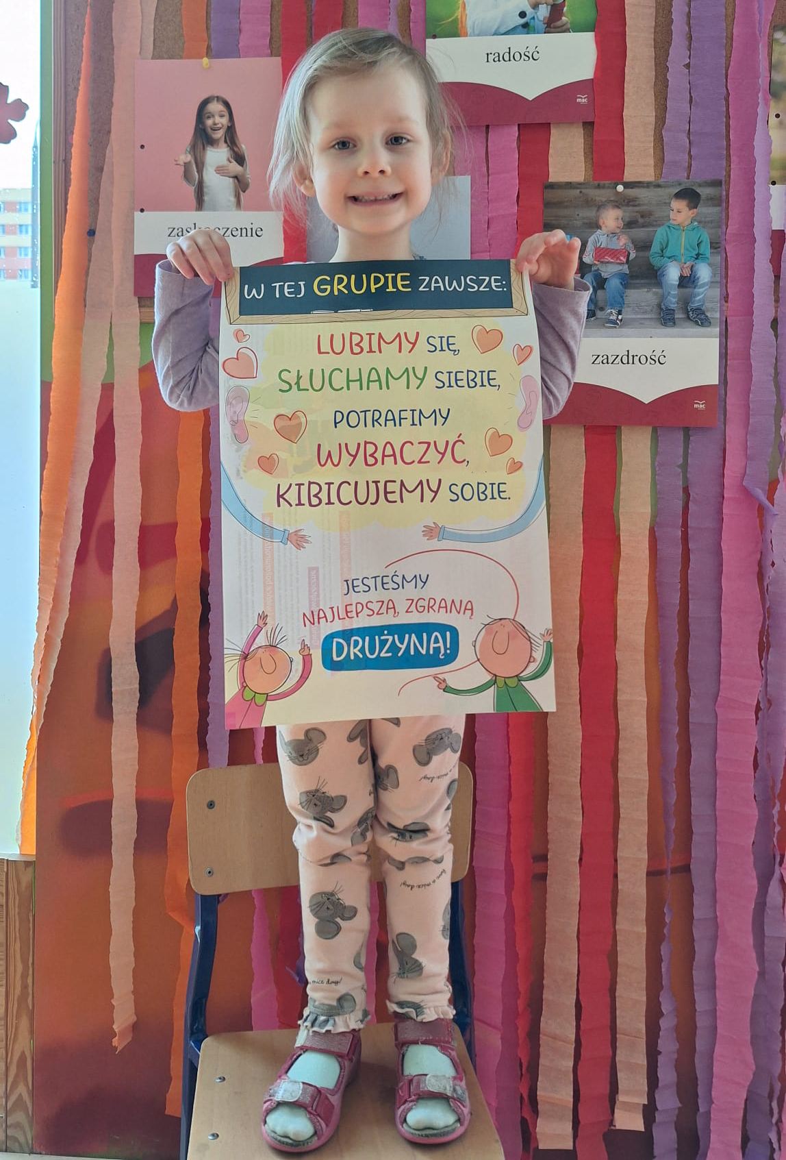 Dziecko w przedszkolu prezentuje plakat z zasadami grupy, podkreślającymi współpracę, wzajemny szacunek i wsparcie.
